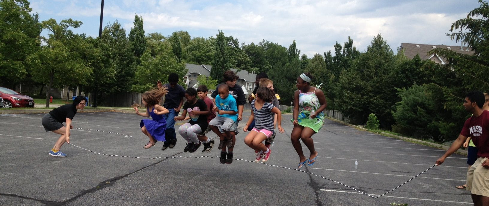 Group of people playing jump rope in a parking lot with trees and buildings in the background.